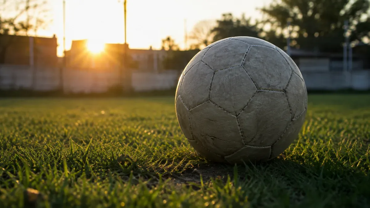 Primer plano de una pelota de fútbol sobre el césped de una cancha de barrio, en contraluz de tarde.